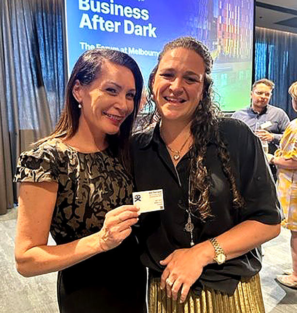 Two women posing together at an event with a screen displaying 'Business After Dark' in the background, with one woman holding a braille business card.