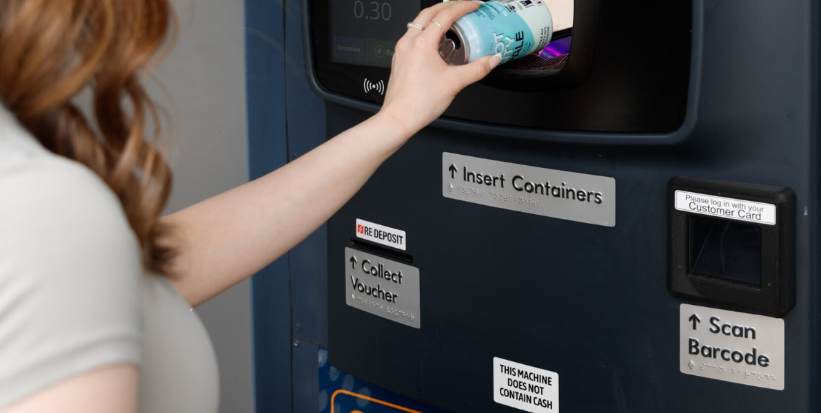 Person using a recycling machine labeled 'VIST' to recycle items, with braille signs for machine usage instructions.