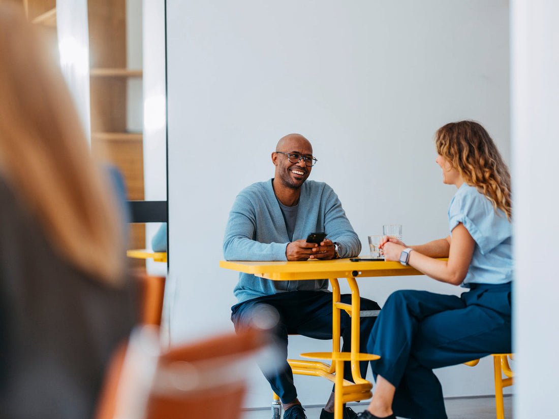 A man and a woman are seated at a bright yellow table in a modern indoor setting, smiling and talking. The man wears a light blue cardigan over a gray shirt, and the woman wears a light blue top and dark pants.