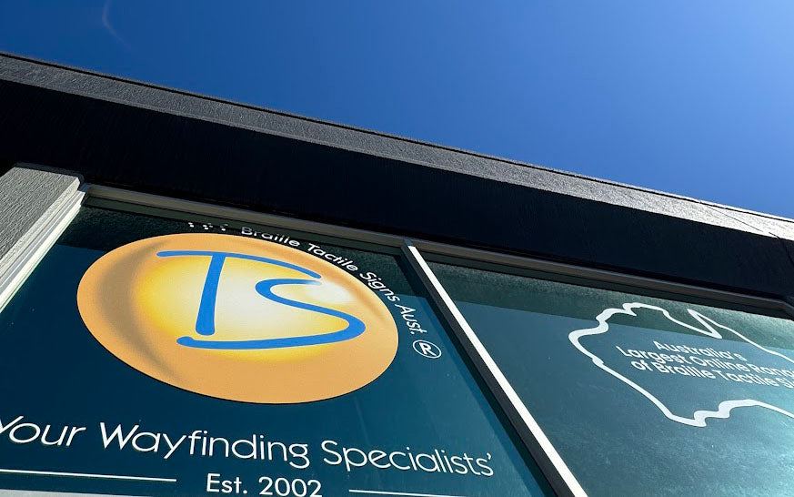 Braille Tactile Signs logo on a window sign with 'Your Wayfinding Specialists' text against a blue sky.