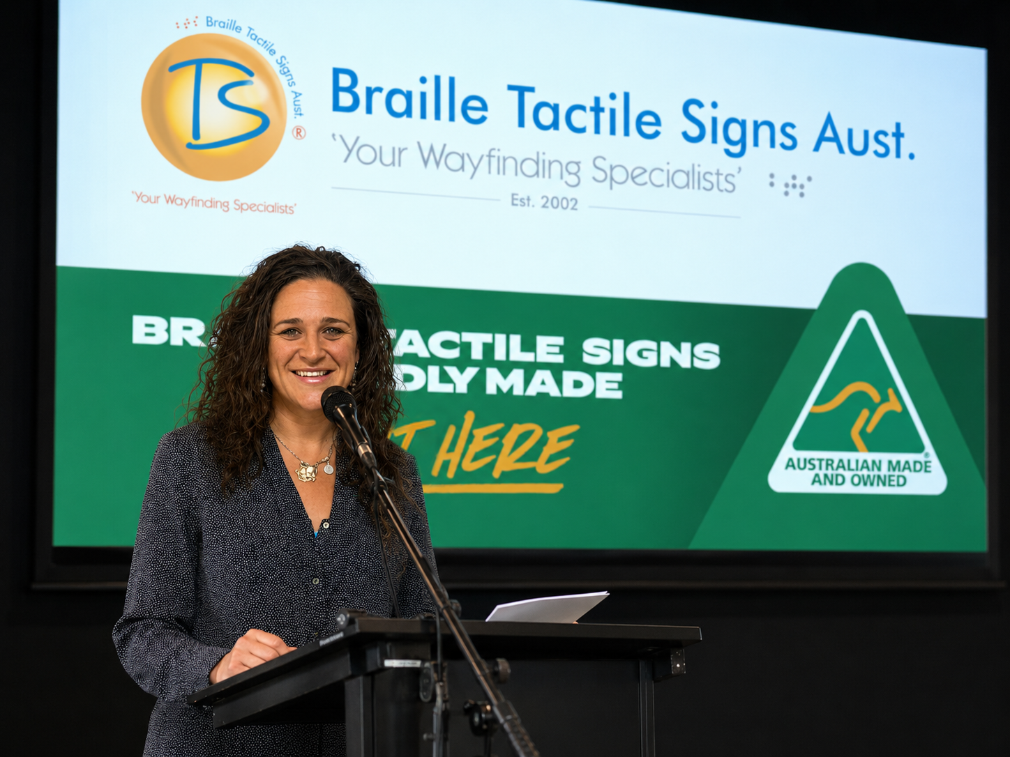 Claudia Gatt speaking at a podium with Braille Tactile Signs Aust. branding on a screen behind her.