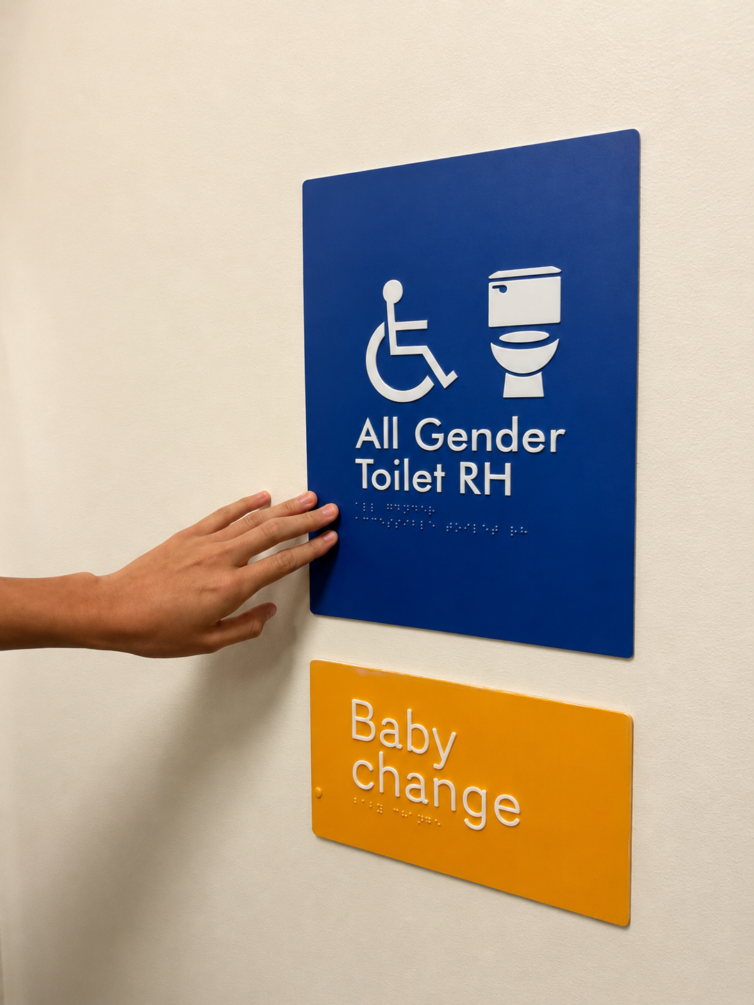 Person touching a Braille tactile sign reading "All Gender Toilet LH" in raised tactile text with corresponding Braille beneath. A second sign below reads "Baby Change" in raised tactile text with corresponding Braille.