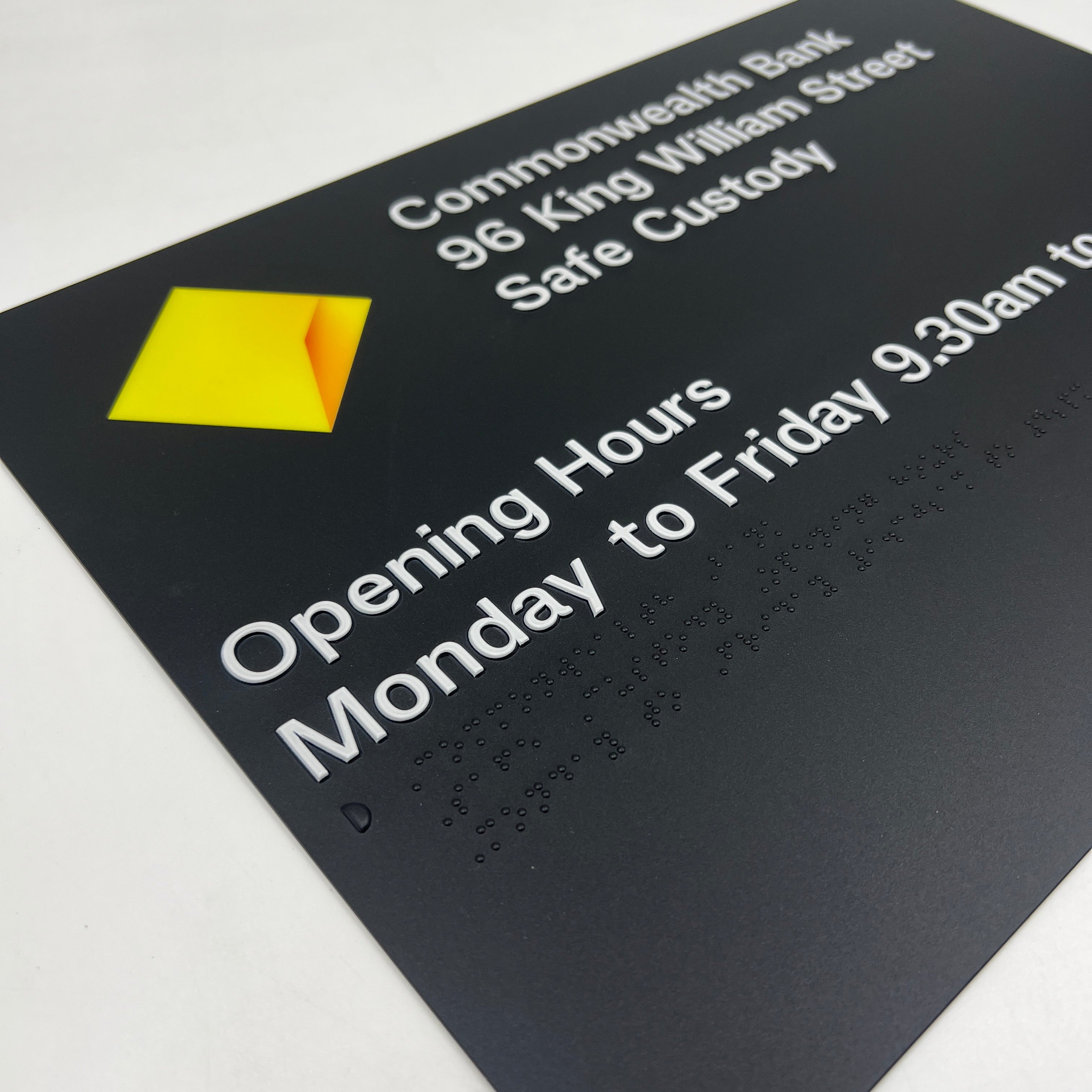Black braille sign with Commonwealth Bank logo and opening hour text.