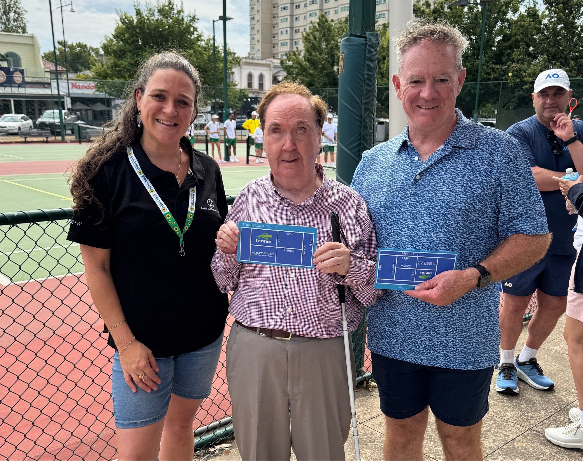 Two men and a woman holding blue tactile maps of a tennis court, standing infront of a tennis court, with people in the background.