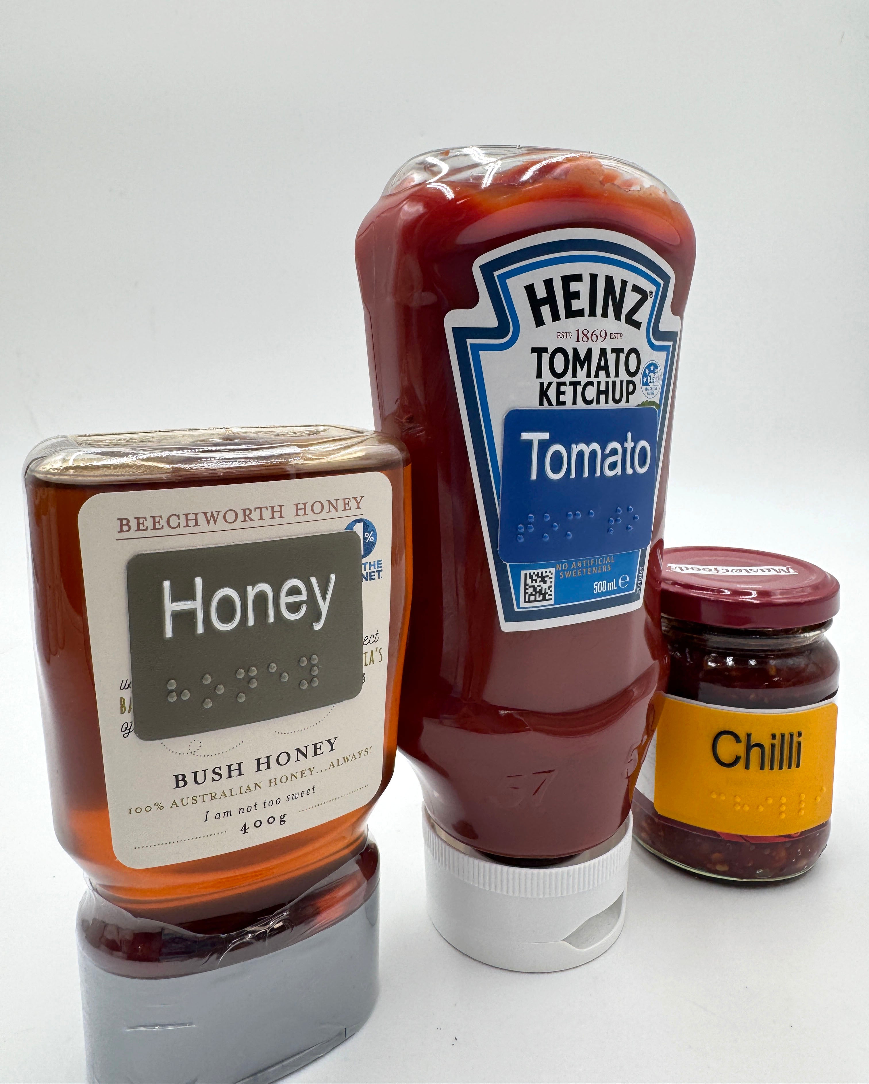 Three condiment bottles: a jar of honey, a bottle of Heinz tomato ketchup, and a jar of chilli, all with braille labels on them.