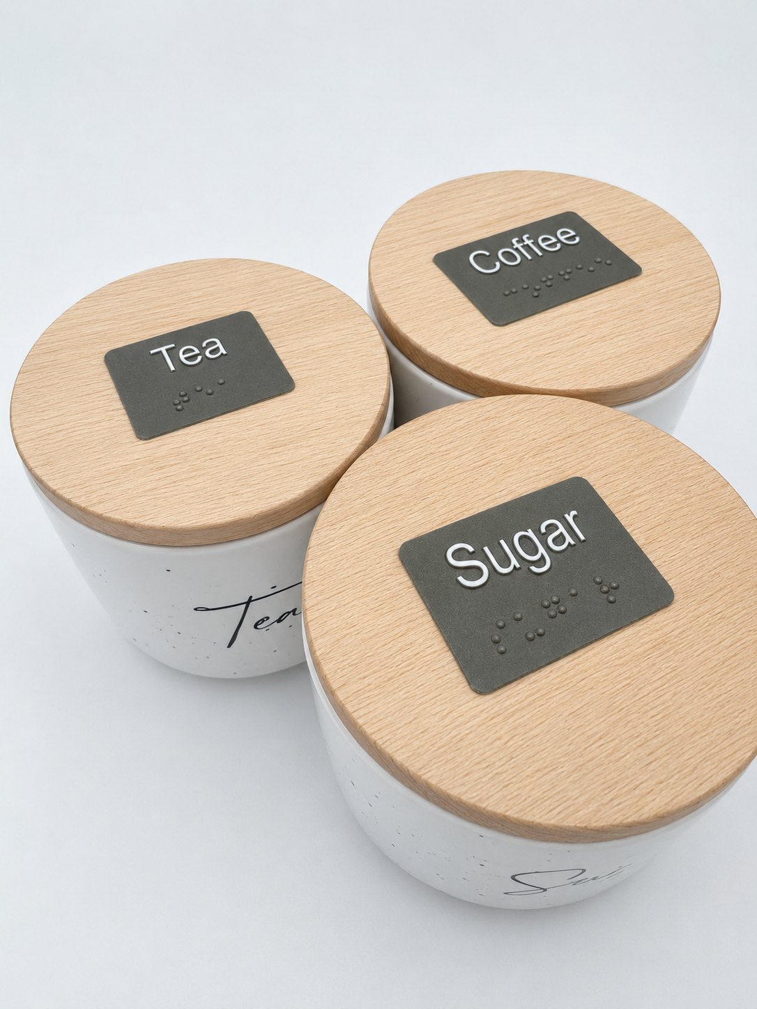Three white containers with wooden lids with braille labels 'Tea', 'Coffee', and 'Sugar' on a light gray background.