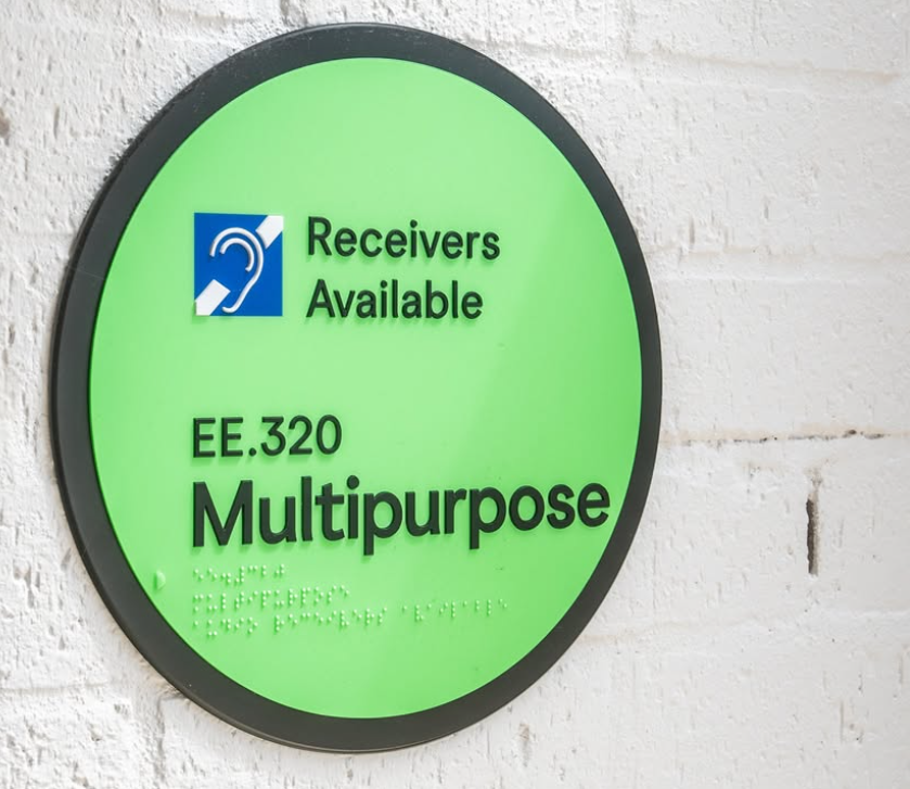 Circular green sign with text, braille and a logo on a white brick wall background